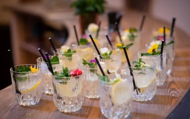 Row of clear cocktails with black straws garnished with colorful edible flowers and fresh herbs